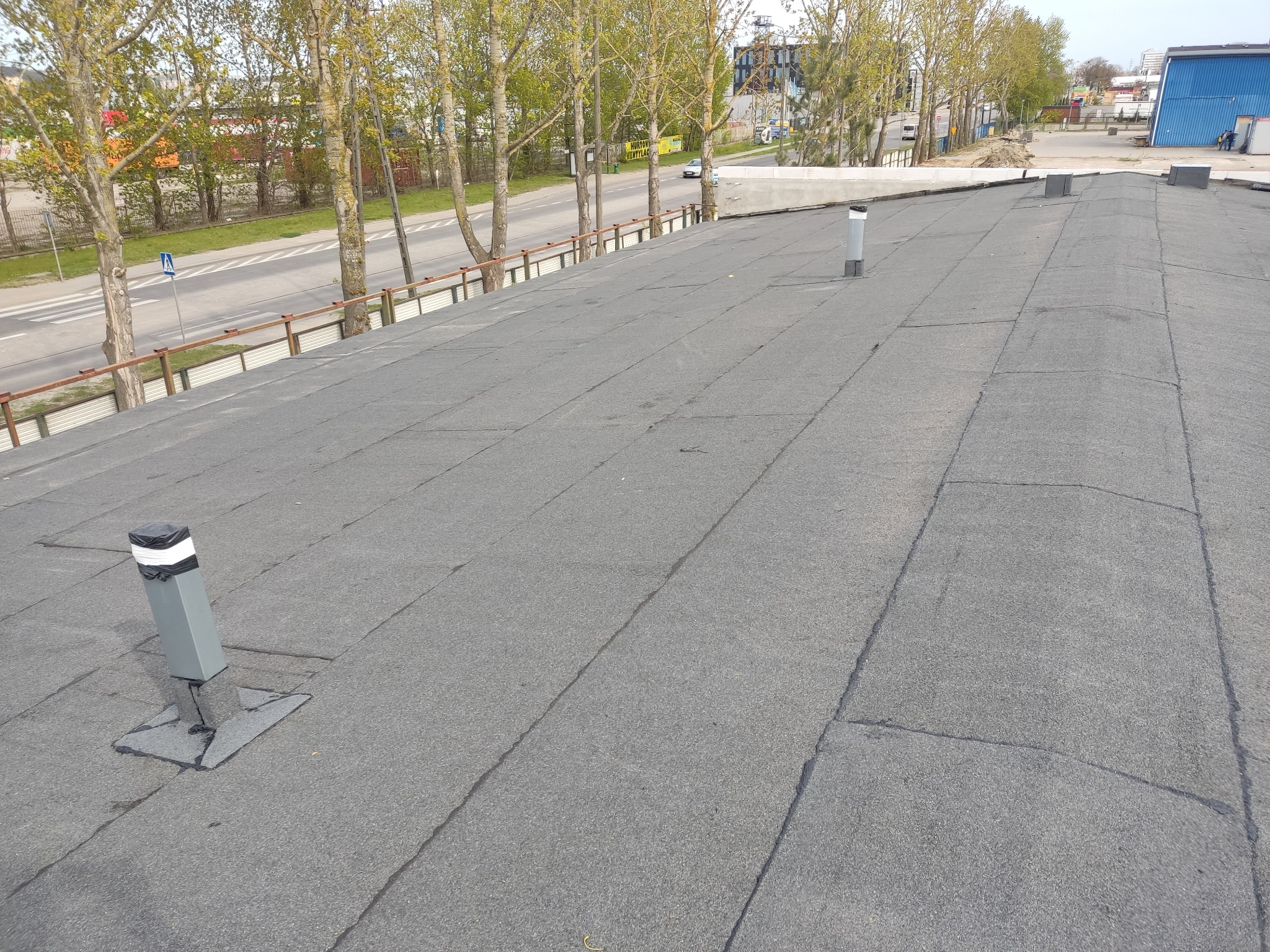 Płaski dach pokryty szarą papą bitumiczną, widoczne łączenia i kominki wentylacyjne, w tle ulica z drzewami i budynki.