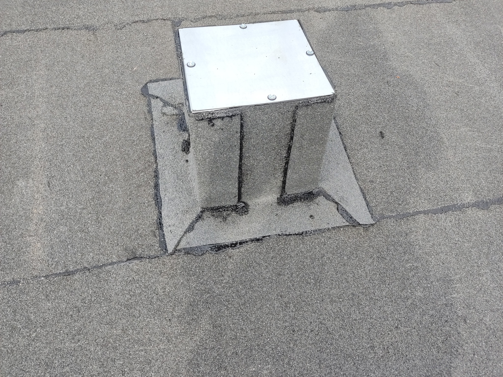 Płaski dach pokryty papą z metalową obudową kominka wentylacyjnego lub innego elementu, zabezpieczoną czterema śrubami.