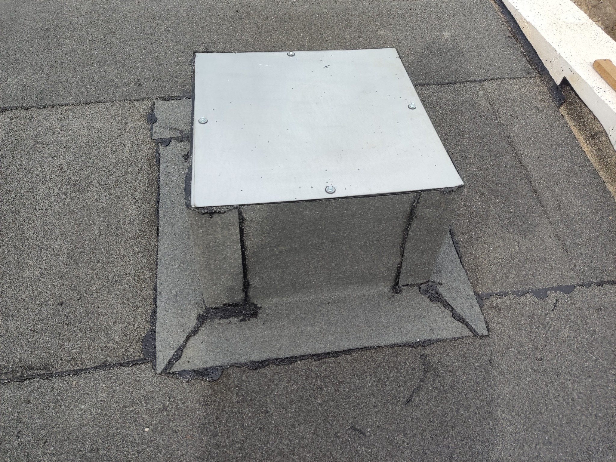 Szczelne przejście wentylacyjne na dachu pokrytym papą, zabezpieczone metalową osłoną z czterema śrubami.