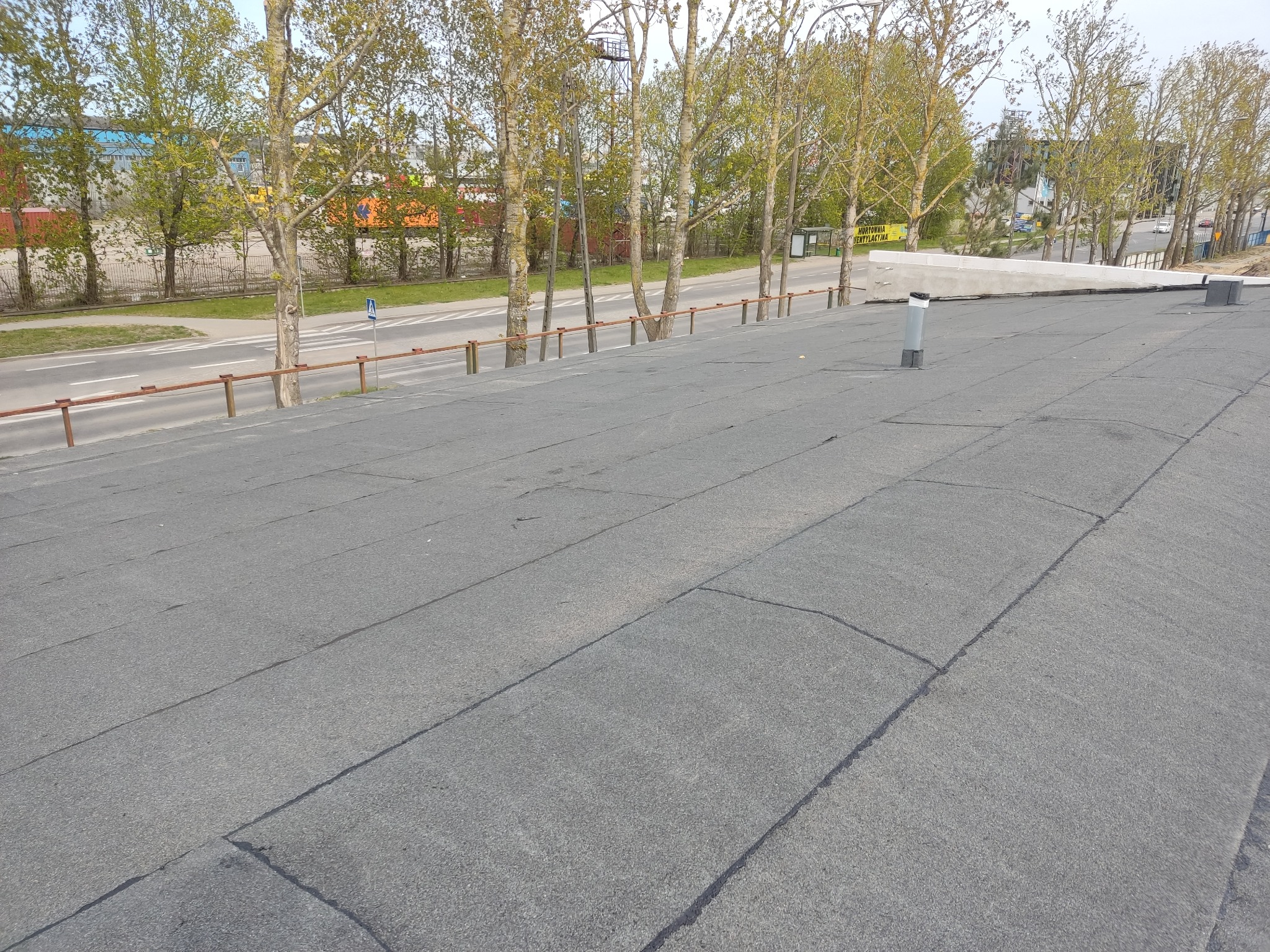 Płaski dach pokryty papą termozgrzewalną, widoczne łączenia i kominek wentylacyjny, w tle rząd drzew i budynki przemysłowe.