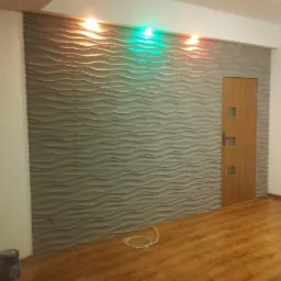 Szara ściana z panelami 3D o falistej strukturze, podświetlona od g&oacute;ry pomarańczowym i zielonym światłem, obok drewniane drzwi z przeszkleniami, na podłodze panele imitujące drewno.