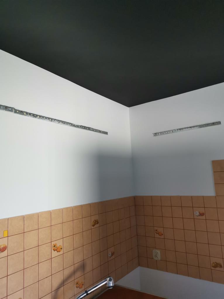 Przygotowana ściana do montażu szafek kuchennych: białe ściany, czarny sufit, zamontowane szyny montażowe, stara glazura w dolnej części ściany.