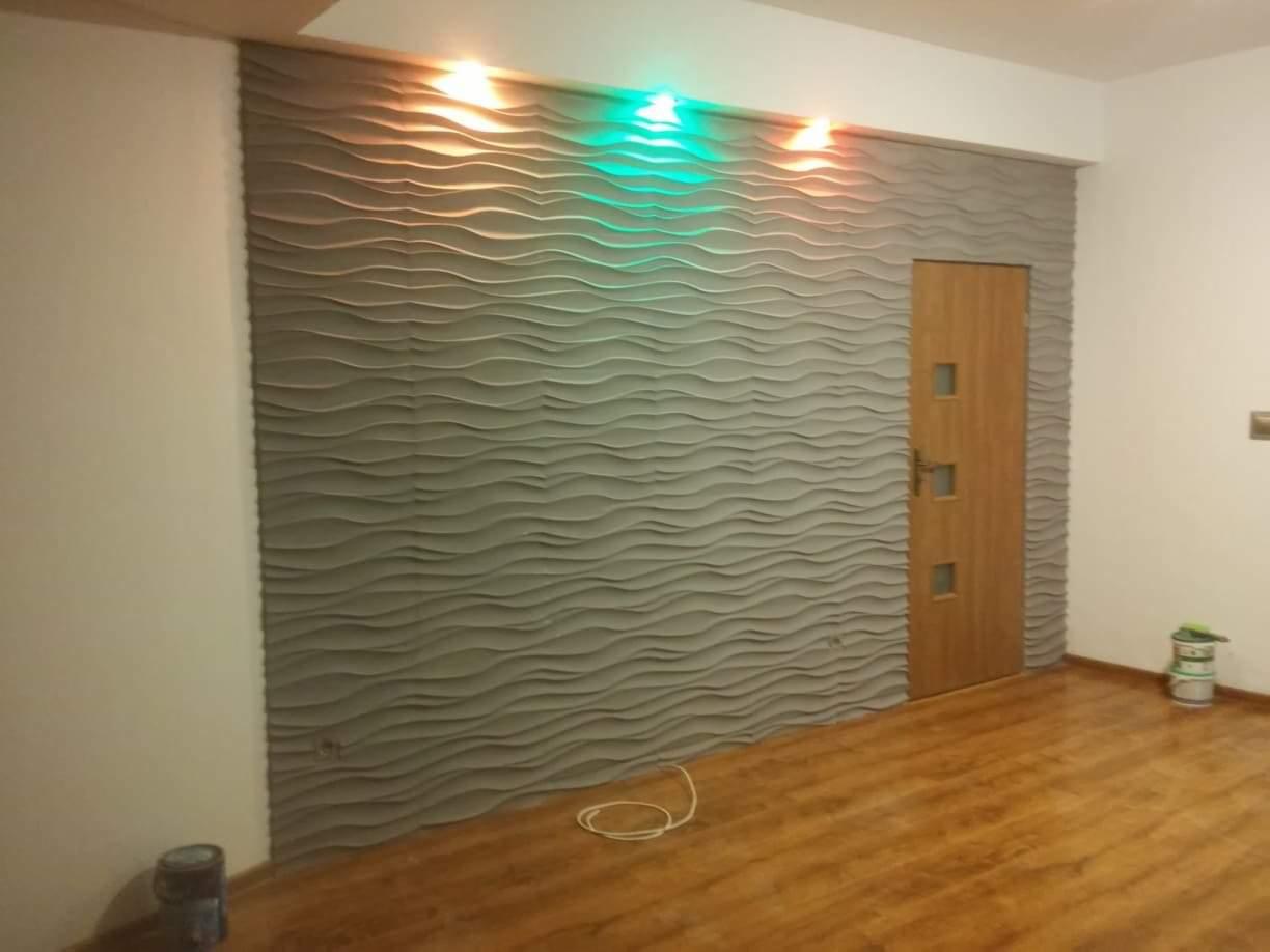 Szara ściana z panelami 3D o falistej strukturze, podświetlona od góry pomarańczowym i zielonym światłem, obok drewniane drzwi z przeszkleniami, na podłodze panele imitujące drewno.