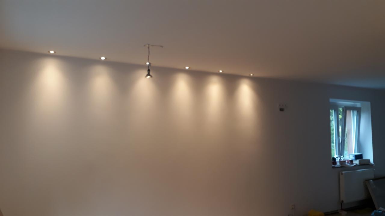 Świeżo pomalowane na biało wnętrze z zamontowanymi reflektorami w suficie i wiszącą żarówką, widok na okno z firanką i grzejnik.