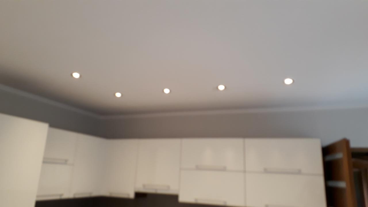 Biały sufit z rzędem sześciu punktowych lamp halogenowych w nowocześnie urządzonej kuchni z białymi szafkami.