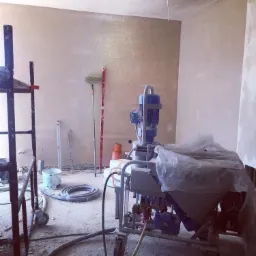 Wnętrze w trakcie prac tynkarskich: ściany i sufit pokryte świeżym tynkiem, na podłodze maszyna do tynkowania pod folią ochronną, obok rusztowanie i akcesoria malarskie.