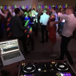 Parkiet taneczny w trakcie imprezy, widoczny sprzęt DJ-ski - laptop i konsoleta, w tle tańczący ludzie pod kolorowymi światłami.