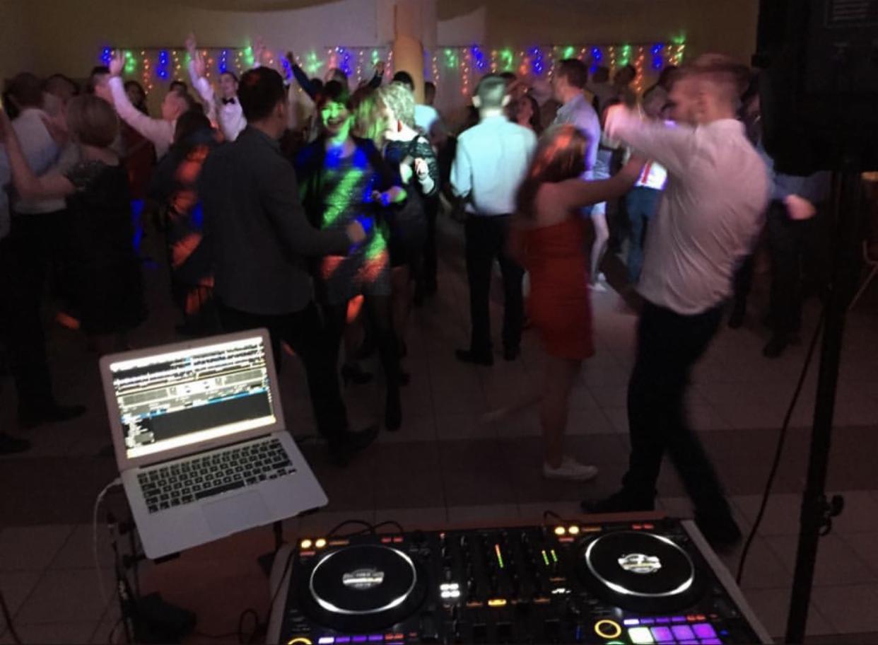 Parkiet taneczny w trakcie imprezy, widoczny sprzęt DJ-ski - laptop i konsoleta, w tle tańczący ludzie pod kolorowymi światłami.