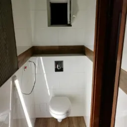 Wykończona łazienka z wiszącą toaletą, białymi płytkami i drewnopodobną podłogą oraz obramowaniem, okno z otwartym skrzydłem.