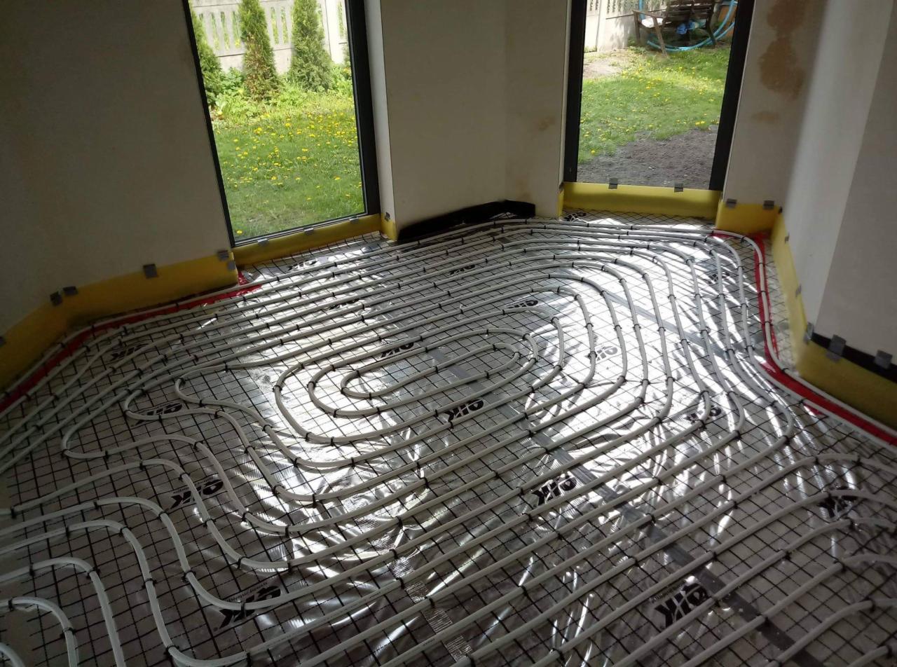 Białe rury ogrzewania podłogowego ułożone na srebrnej folii z czarną siatką, widoczne żółte paski pianki dylatacyjnej przy ścianach i okna z widokiem na ogród