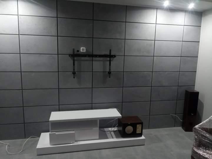 Ściana wykończona płytami imitującymi beton, z zamontowanym uchwytem na telewizor, poniżej biała szafka RTV i głośniki.