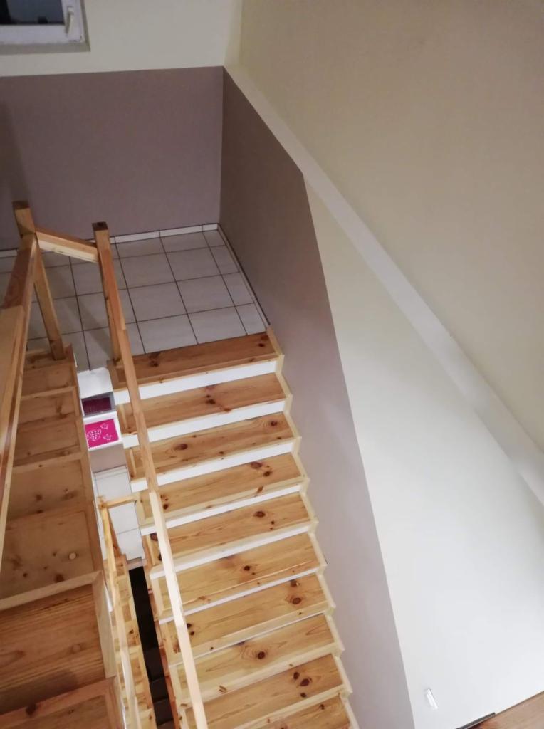 Schody drewniane z surowego drewna z białymi podstopnicami, widok z góry na tle pomalowanych ścian i płytek podłogowych.