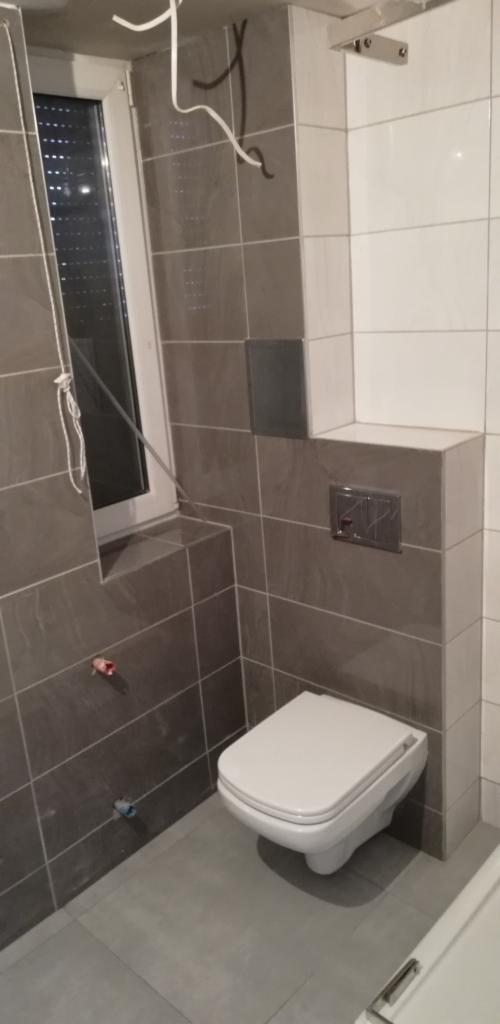 Świeżo wyremontowana łazienka w Zabrzu z szarymi i białymi płytkami, wiszącą toaletą i ukrytą spłuczką. Widoczne wystające kable i rurki przed zakończeniem prac.