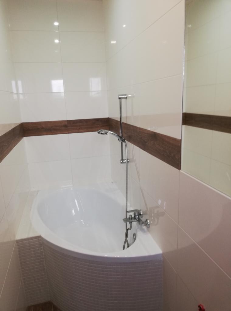 Nowoczesna łazienka z białą, asymetryczną wanną obudowaną mozaiką, chromowaną armaturą i białymi płytkami z dekoracyjnym pasem imitującym drewno.