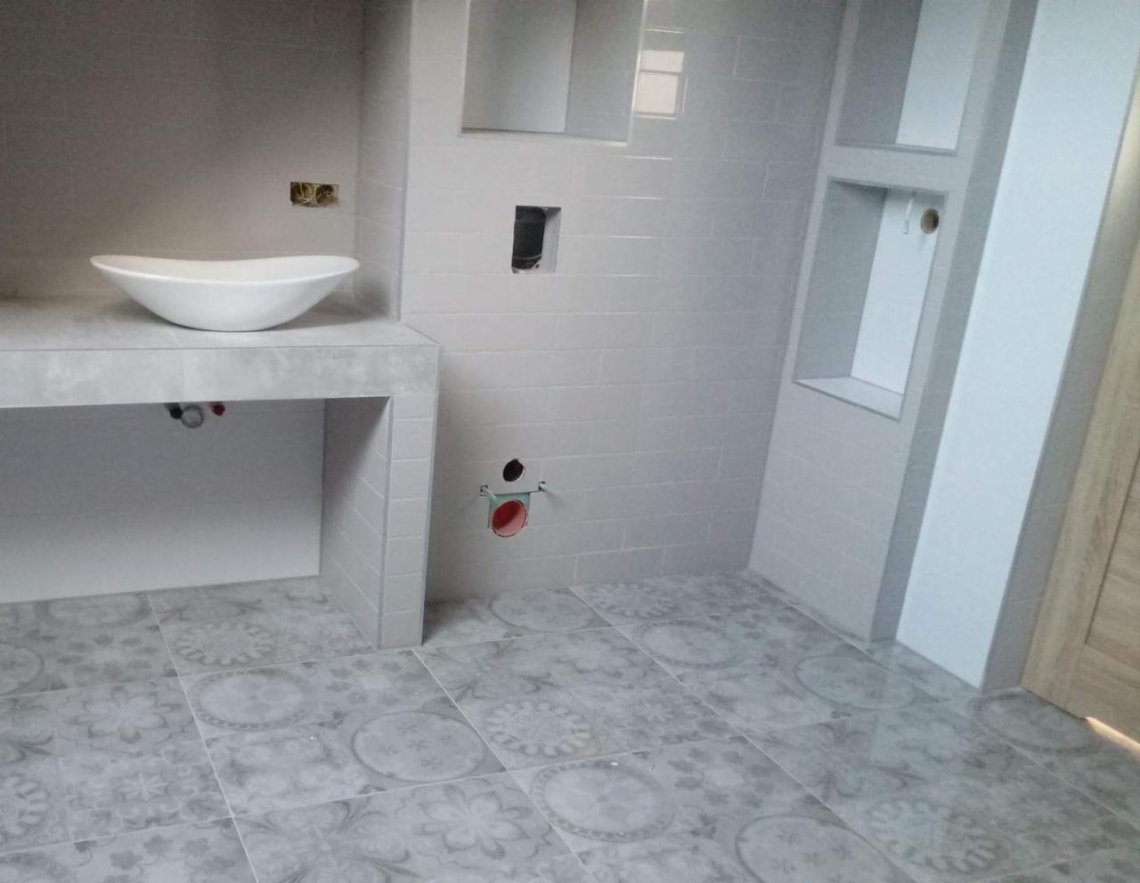 Nowoczesna łazienka w trakcie wykończenia: biała umywalka nablatowa, szare płytki z dekoracyjnym wzorem na podłodze, widoczne otwory instalacyjne w ścianie z szarych płytek.