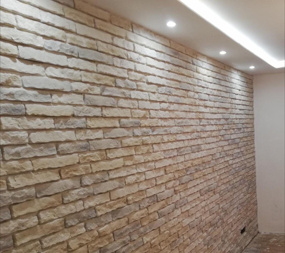 Ściana wykończona dekoracyjną okładziną imitującą kamień w odcieniach beżu i szarości, z podświetleniem sufitowym punktowym i liniowym.