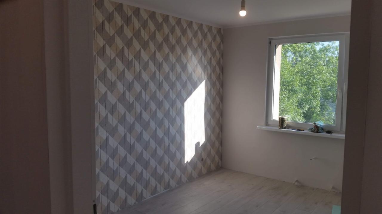 Pokój po remoncie z wzorzystą tapetą na ścianie, drewnianą podłogą i oknem z widokiem na zieleń, na parapecie leżą narzędzia i materiały budowlane.