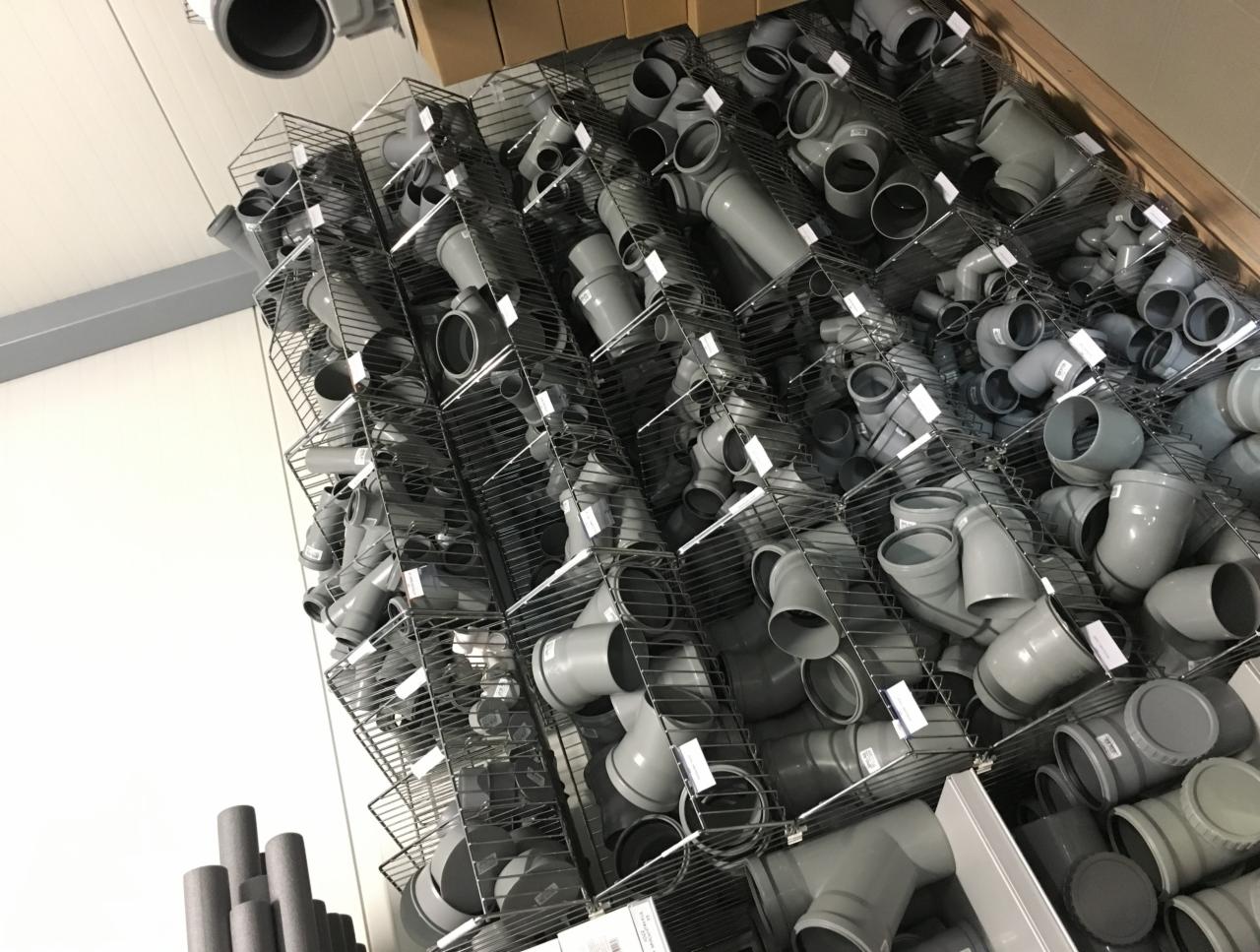 Regał sklepowy wypełniony szarymi kształtkami i rurami PCV, widoczne kolanka, trójniki i proste odcinki, ułożone w metalowych koszach.