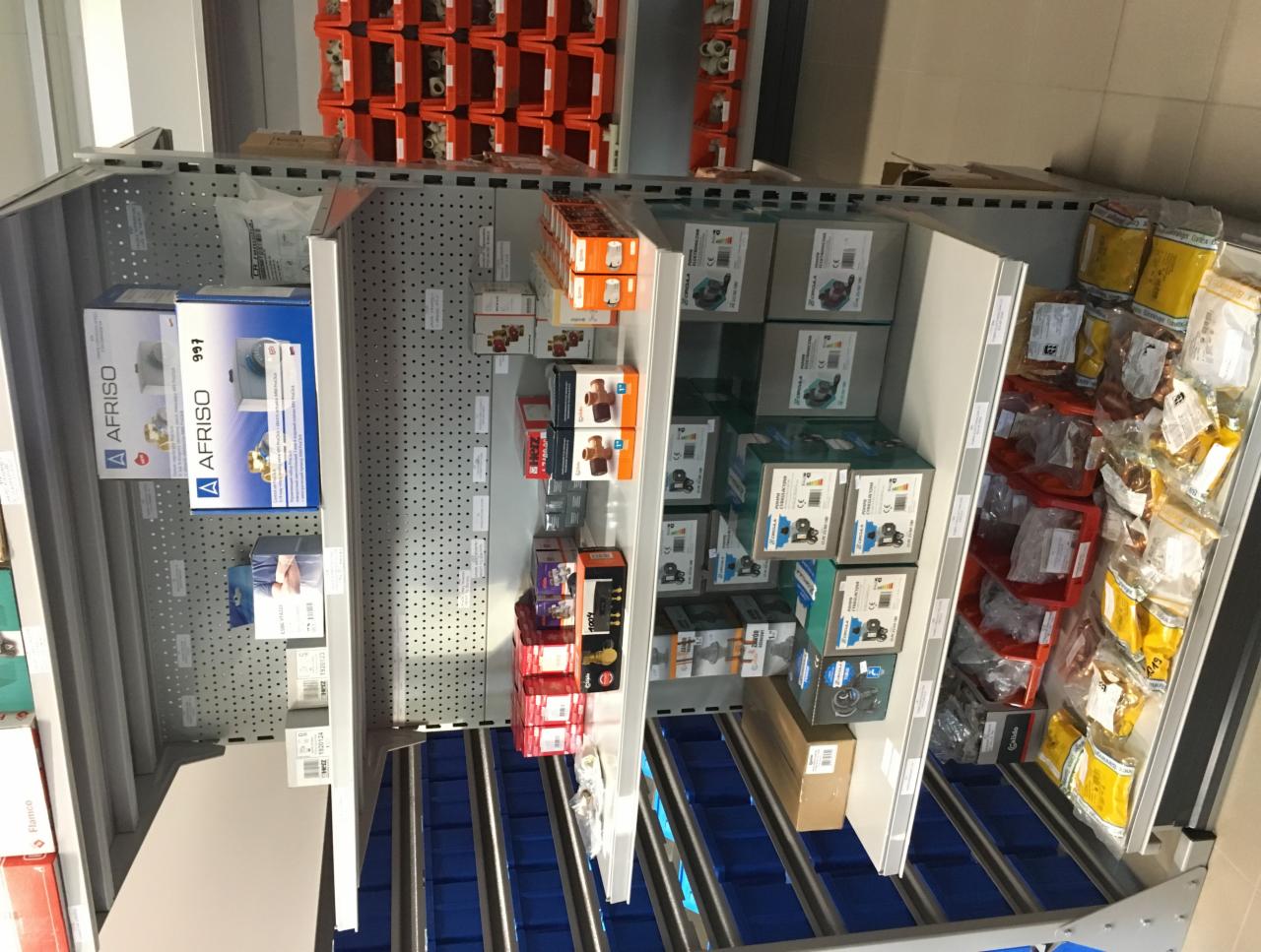 Półki magazynowe z zapasem komponentów i akcesoriów AFRISO do instalacji pomp ciepła, widoczne opakowania zbiorcze i pojedyncze elementy w opakowaniach foliowych.