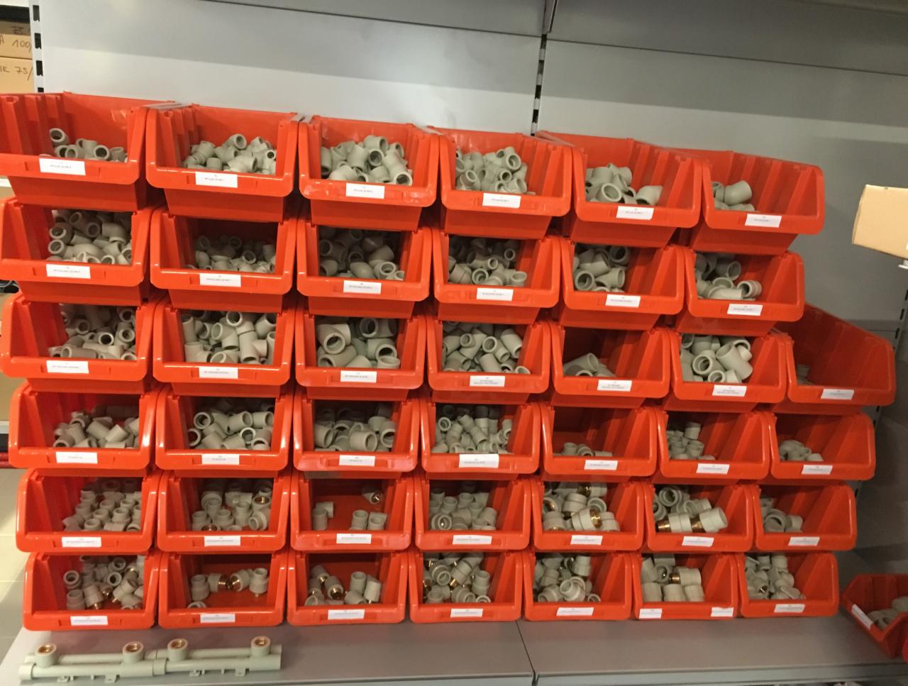 Regał z pomarańczowymi pojemnikami wypełnionymi złączkami instalacyjnymi z tworzywa sztucznego i mosiądzu.