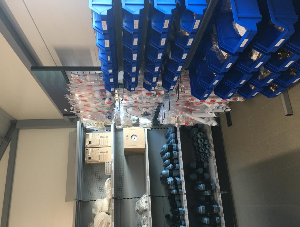 Magazyn z rzędami niebieskich pojemników i półkami wypełnionymi rurami, złączkami i elementami instalacyjnymi w opakowaniach foliowych i kartonach.