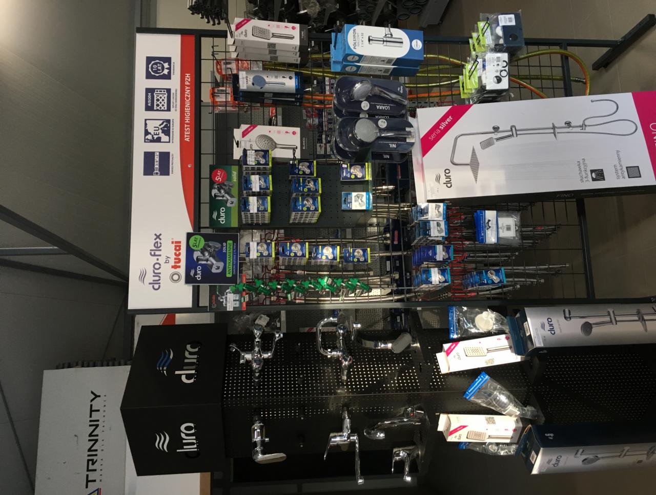 Ekspozycja armatury łazienkowej w sklepie: baterie, słuchawki prysznicowe, zestawy natryskowe, opakowania produktów marki Cluro i Trinity, widoczne logo producentów i atesty higieniczne.
