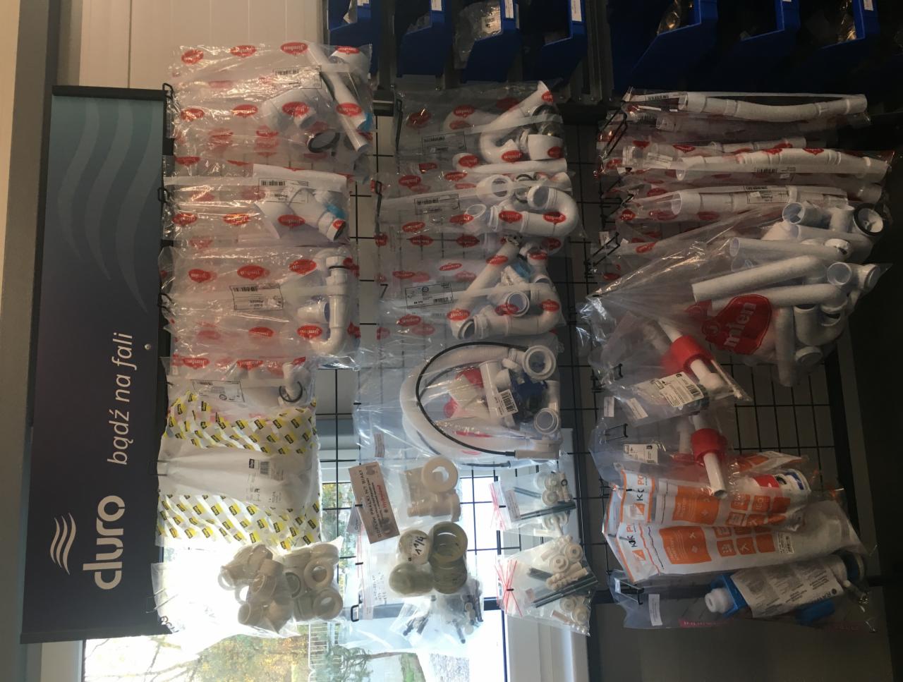 Różne kształty i rozmiary białych rur i złączek hydraulicznych zapakowanych w przezroczyste plastikowe torby, wiszące na metalowej siatce w sklepie, obok reklamy z logo firmy Cluro.