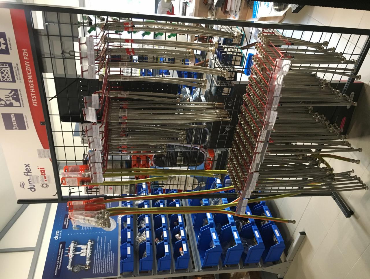 Ekspozycja w sklepie z instalacjami: metalowe węże elastyczne, złączki i rozdzielacze na regale z niebieskimi pojemnikami i materiałami reklamowymi producenta.