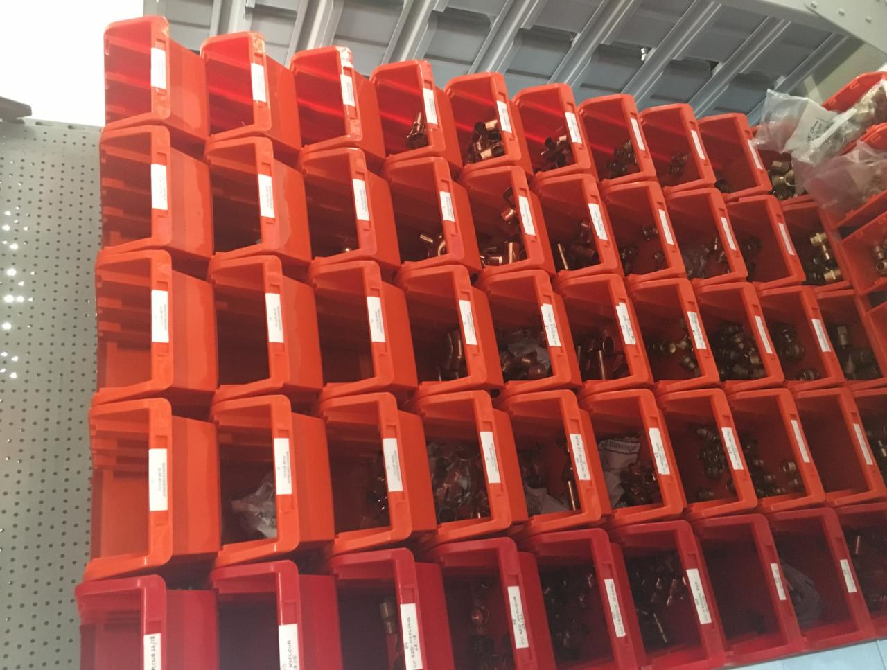 Magazyn komponentów instalacyjnych: rzędy pomarańczowych pojemników z mosiężnymi złączkami, etykiety na każdym pojemniku, metalowa perforowana ściana w tle.