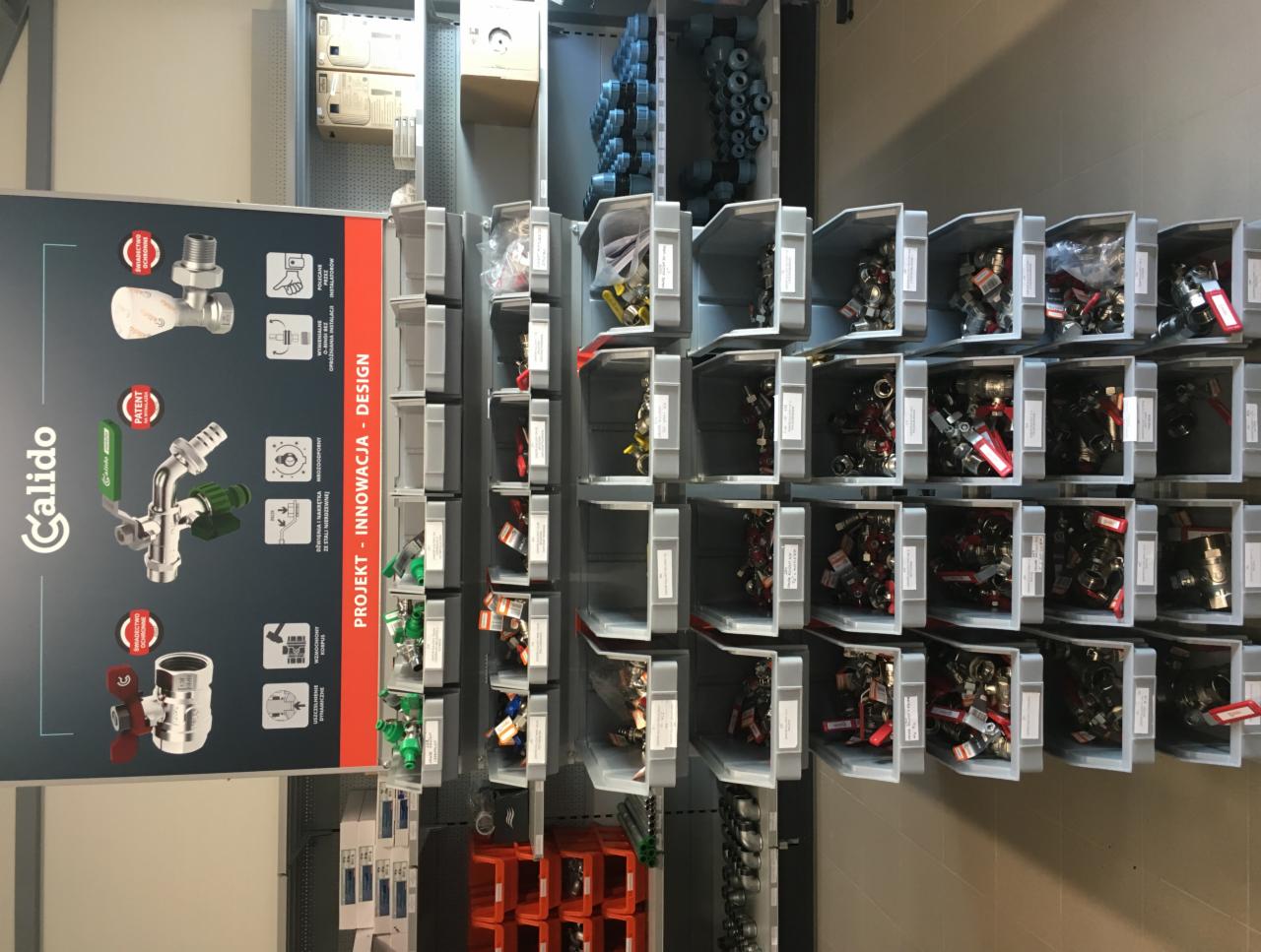 Regał z szufladami zawierającymi złączki hydrauliczne i zawory, obok plansza reklamowa Calido prezentująca produkty.