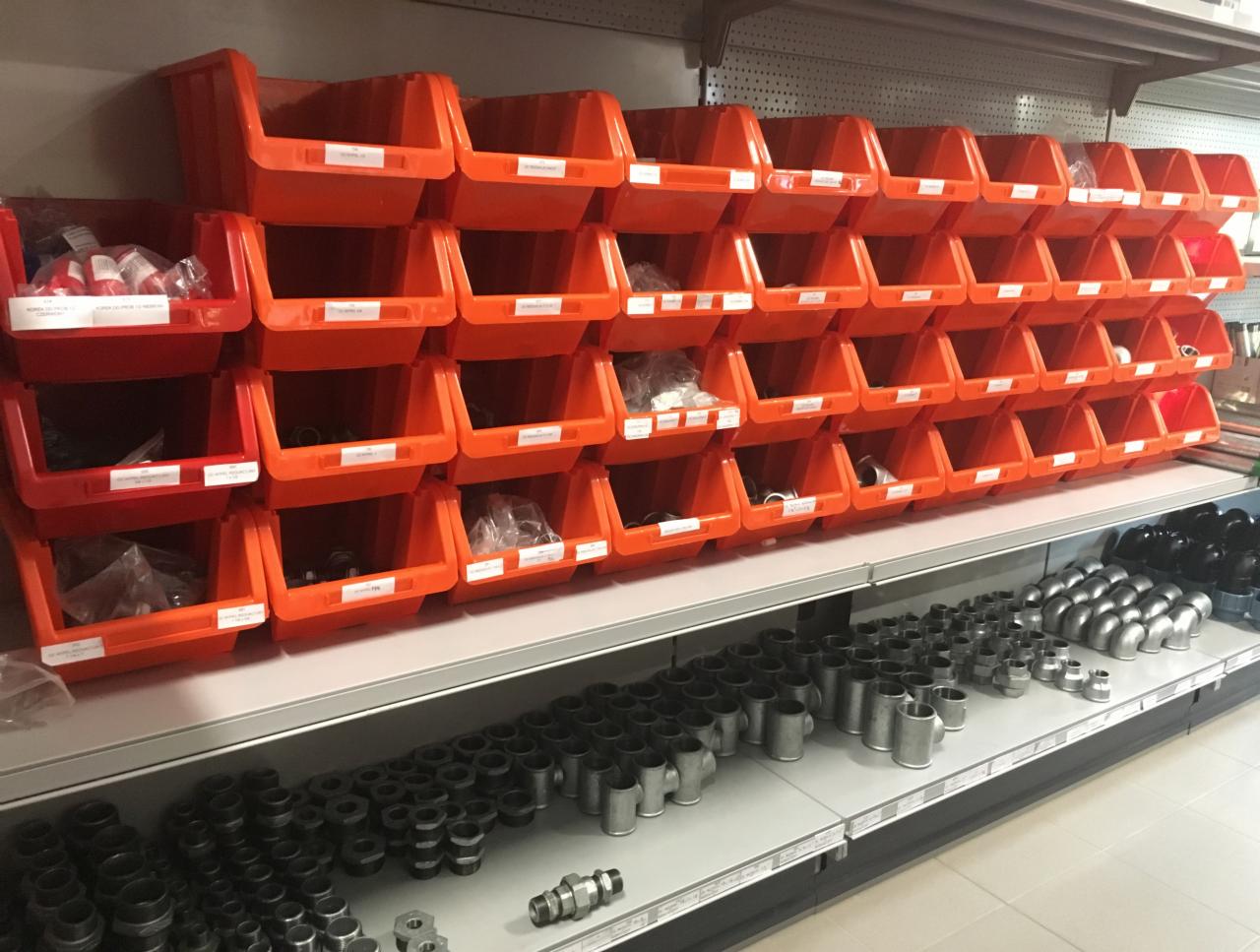 Regał z pomarańczowymi pojemnikami wypełnionymi złączkami i rurami, poniżej na półkach metalowe kształtki i śrubunki.