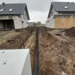 Wykop z ułożonymi niebieskimi rurami kablowymi między dwoma budynkami w stanie surowym, pochmurne niebo.