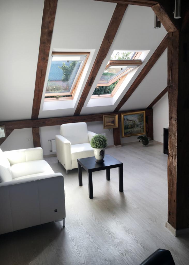 Jasne, przestronne poddasze z drewnianymi belkami, oknami dachowymi wpuszczającymi światło, białymi sofami i stolikiem kawowym z rośliną doniczkową, uzupełnione obrazami na ścianie.