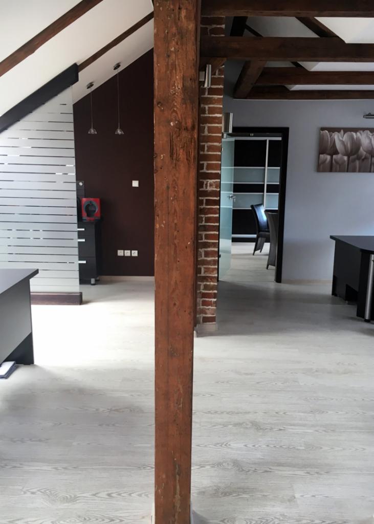 Wnętrze biura z drewnianą kolumną na pierwszym planie, widoczne biurka, przeszklone drzwi i elementy dekoracyjne.
