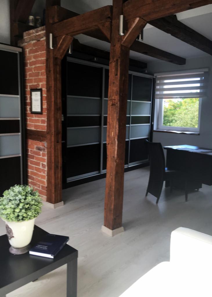 Wnętrze biura z drewnianymi belkami, ceglaną ścianą, zabudową szaf i biurkiem przy oknie z żaluzjami.