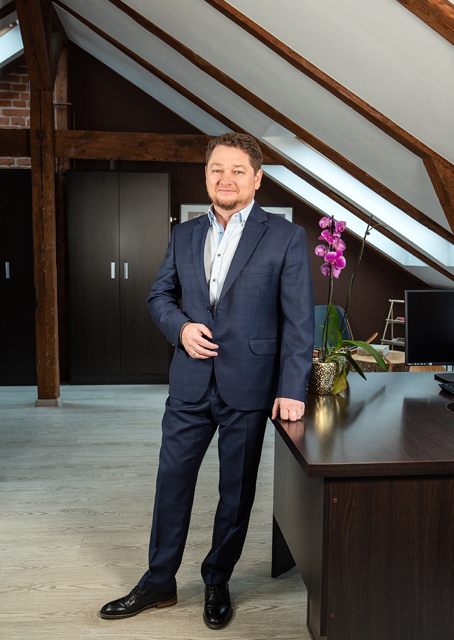 Mężczyzna w granatowym garniturze, stojący w biurze z drewnianą konstrukcją stropu i storczykiem na biurku.