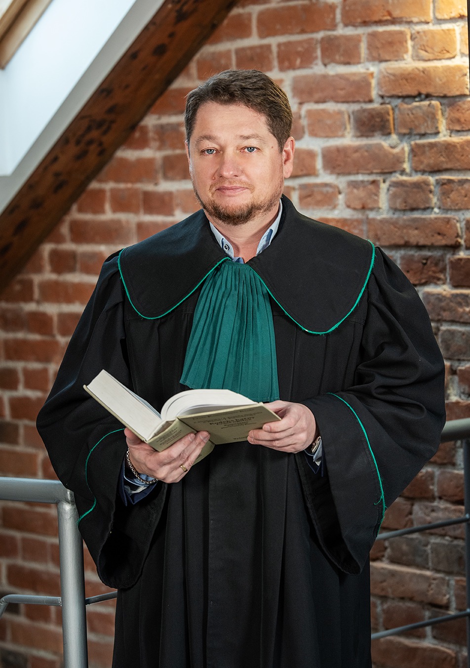 Portret adwokata w todze z zielonym żabotem, trzymającego otwartą książkę na tle ceglanej ściany i drewnianej belki.