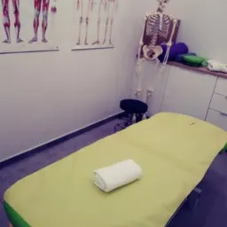 Jasnożółty stół do zabiegów z białym ręcznikiem, w tle plakaty z układem mięśniowym i szkielet stojący obok szafki z poduszkami.