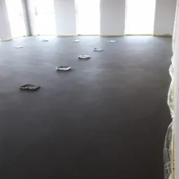 Świeżo wylana, ciemnoszara posadzka jastrychowa w dużym, jasnym pomieszczeniu z widocznymi puszkami instalacyjnymi w podłodze i zabezpieczonymi folią ścianami.