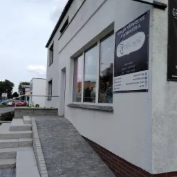 Wejście do budynku z usługami krawieckimi i pasmanterią, z rampą dla niepełnosprawnych. Widoczny szyld z nazwą i godzinami otwarcia.