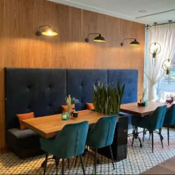 Wnętrze restauracji z drewnianymi blatami stołów, tapicerowanymi krzesłami w kolorze morskim, wysokim, granatowym panelem tapicerowanym wzdłuż ściany i drewnianą boazerią, oświetlone trzema...