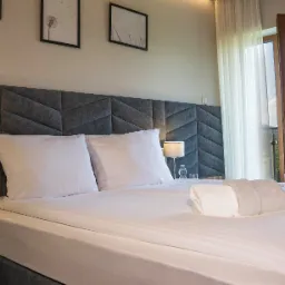 Nowoczesna sypialnia z łóżkiem małżeńskim, szarym tapicerowanym zagłówkiem w geometryczny wzór, białymi poduszkami i pościelą, lampkami nocnymi i otwartym balkonem z widokiem na zieleń.