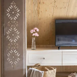 Umeblowanie w pokoju z drewnianą boazerią: szafa z tapicerowanymi drzwiami zdobionymi geometrycznym wzorem, biała komoda z telewizorem, kosz wiklinowy z kocem i różowe kwiaty w wazonie.