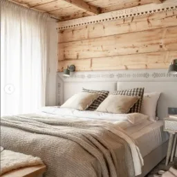 Drewniana sypialnia w stylu rustykalnym z łóżkiem, drewnianym sufitem i ścianami z desek, zdobiona białymi, wycinanymi elementami dekoracyjnymi pod sufitem i na wezgłowiu łóżka.
