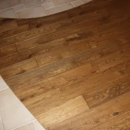 Połączenie podłogi z ciemnego drewna ułożonej w deski z jasnymi, kwadratowymi płytkami ceramicznymi. Zakrzywiona linia podziału materiałów.