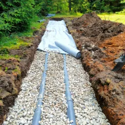Wykop z ułożonymi rurami drenażowymi obsypanymi drobnymi kamieniami, przykryty częściowo białą geowłókniną, widoczne ślady prac ziemnych i fragment łopaty, w tle drzewa iglaste.