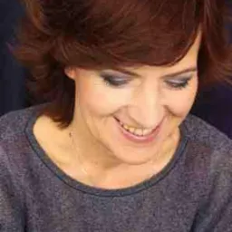 Portret uśmiechniętej kobiety z krótkimi, rudymi włosami, ciemnym makijażem oczu i szarym swetrze na ciemnym tle.