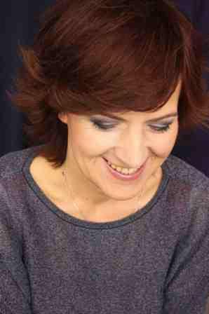 Portret uśmiechniętej kobiety z krótkimi, rudymi włosami, ciemnym makijażem oczu i szarym swetrze na ciemnym tle.