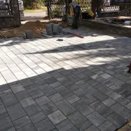 Układanie szarej kostki brukowej na posesji, widoczny pracownik, stos kostki i poziomica, w tle ogrodzenie i drzewa.