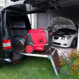 Przenośna, czerwona myjka parowa Optima Steamer umieszczona na wysuwanej platformie z bagażnika czarnego vana, z otwartą maską białego BMW w tle i kwiatami nagietka z przodu.